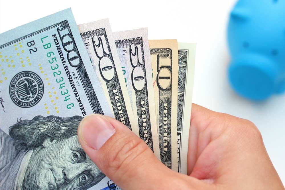 Hand fanning out 100, 50, and 10 dollar bills next to a blue piggy bank.
