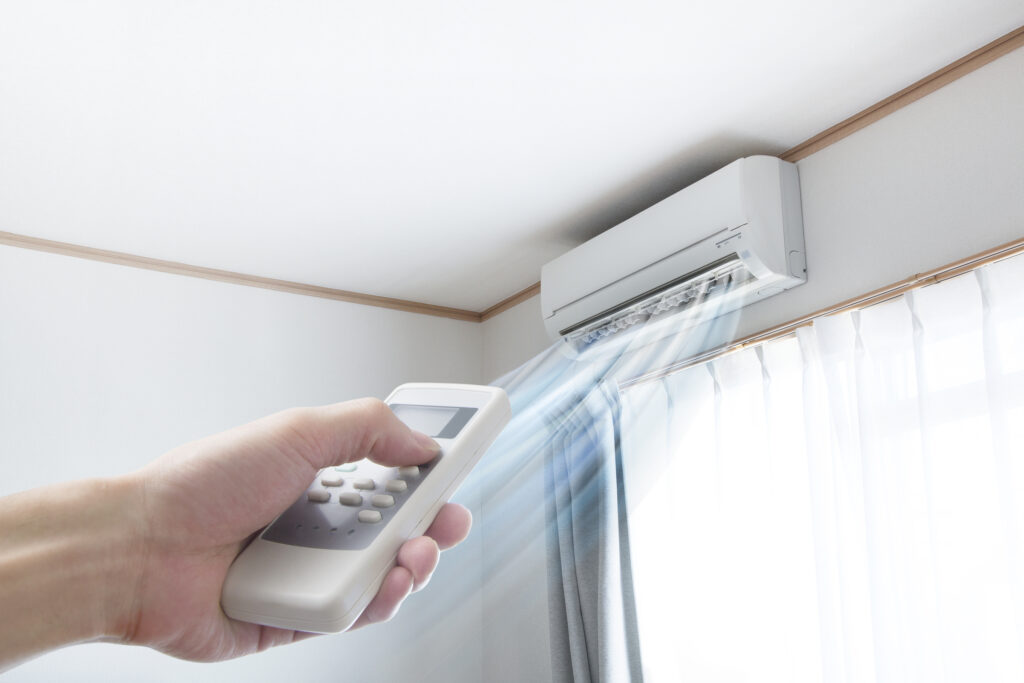 Person's hand aiming a remote at a ductless mini-split installed on a wall, above a window.