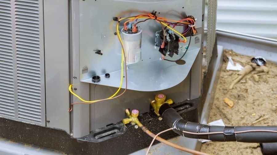 Open access panel of an outdoor air conditioner showing wiring, a capacitor, and exposed electrical components with yellow service valves and insulated refrigerant lines at the base.