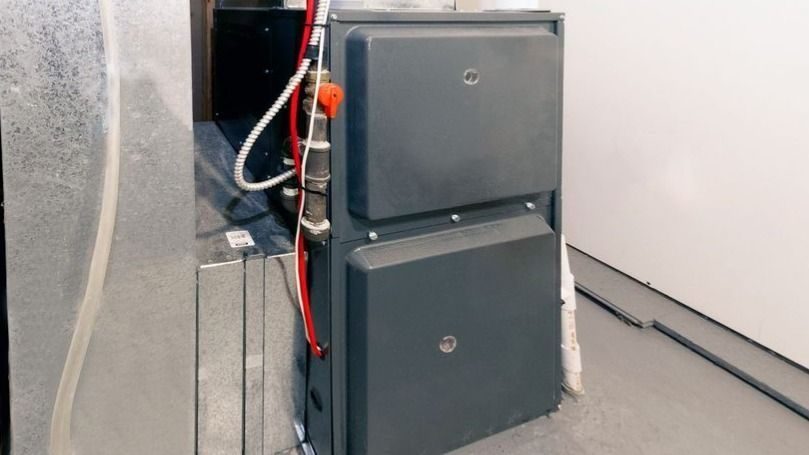 Two gray stacked furnace cabinets in a utility room with exposed ducts and a red gas line.