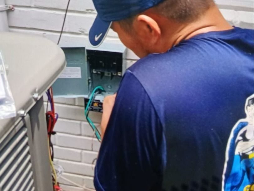 Technician in blue cap and shirt inspecting an outdoor electrical panel of an air conditioner unit outside a building.