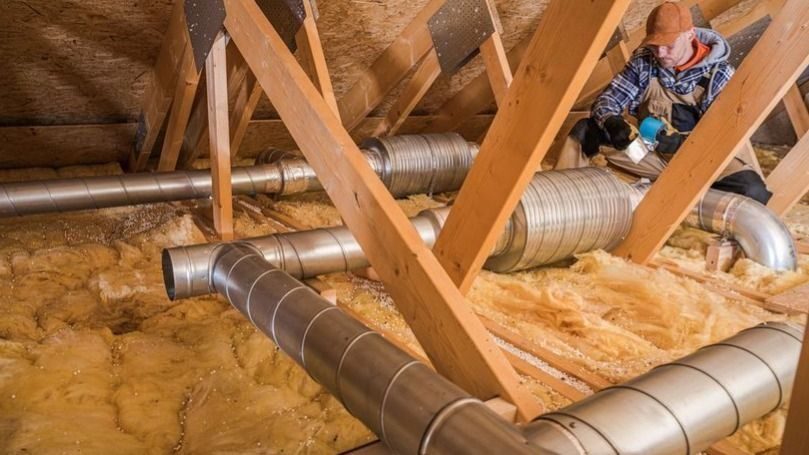 Worker installs or inspects metal HVAC ducts in an attic, surrounded by wooden rafters and fiberglass insulation.