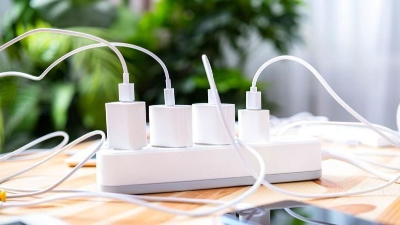 White power strip with multiple plugs and tangled cables on a wooden table, blurred green background.