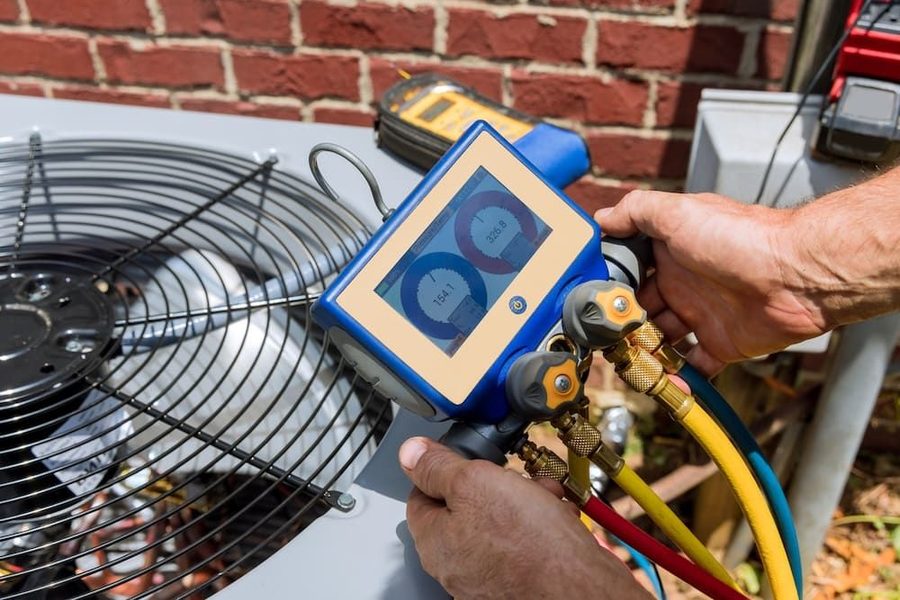 Technician using a refrigerant manifold gauge set on an outdoor AC unit; gauges and colored hoses visible in the setup.