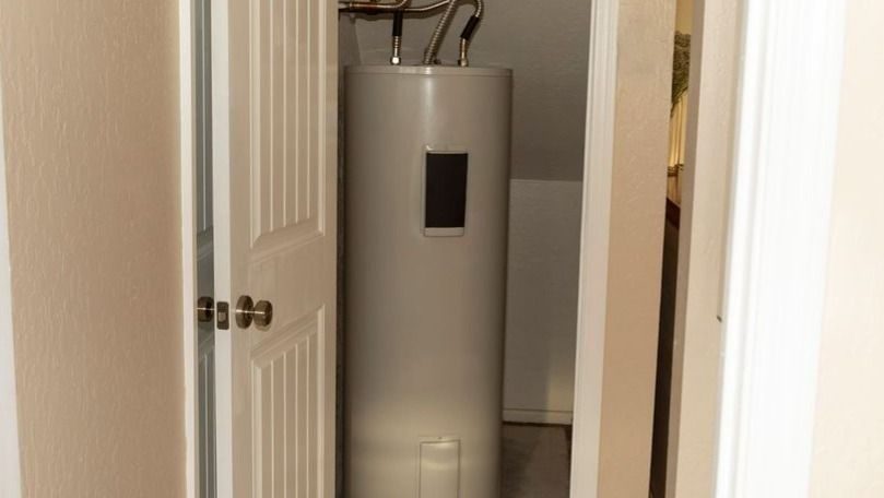 Tall white water heater in a small utility closet with visible pipes at the top, doors ajar on either side of a narrow hallway.